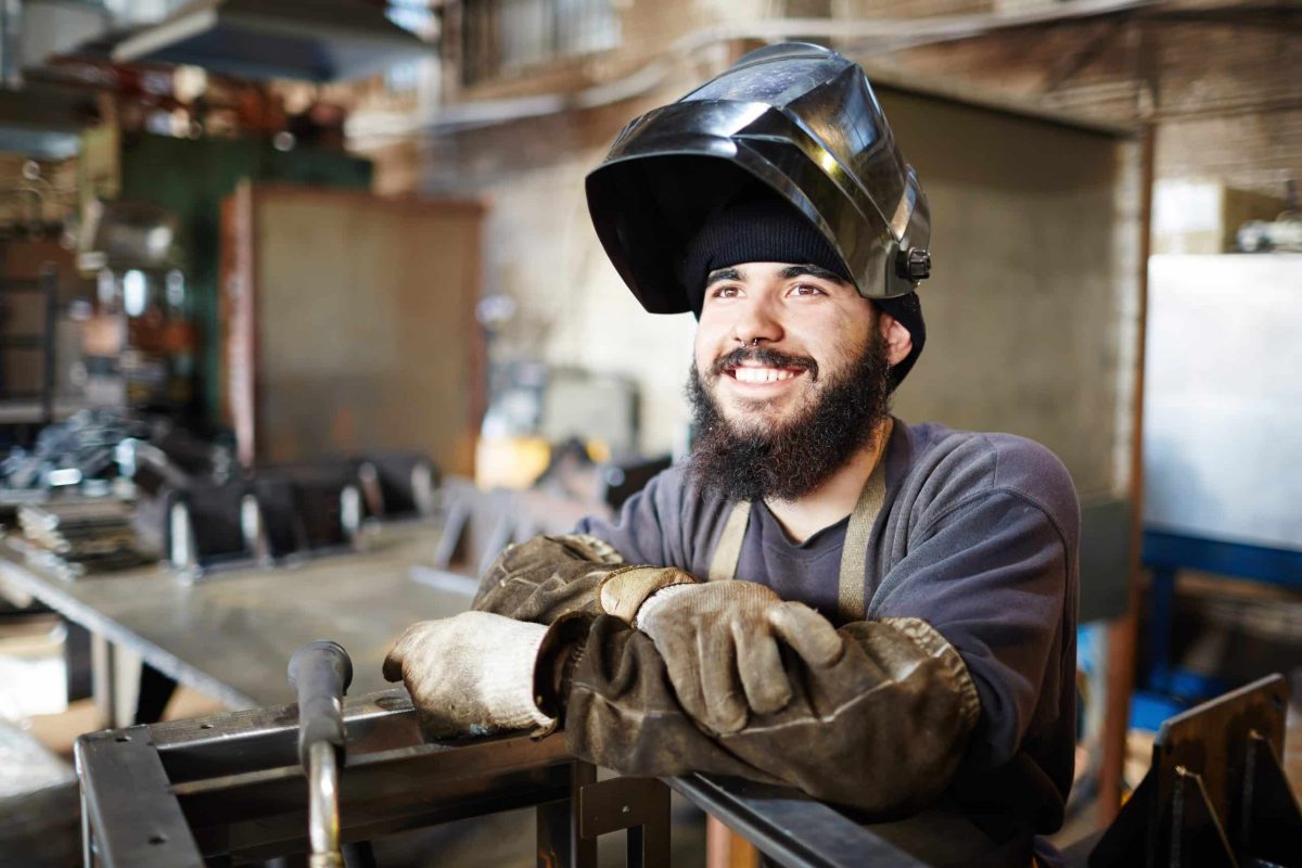 Cheerful dreamy bearded young man with dirty sleeves wearing welding mask and looking with burning eyes while leaning on metal part in factory shop
